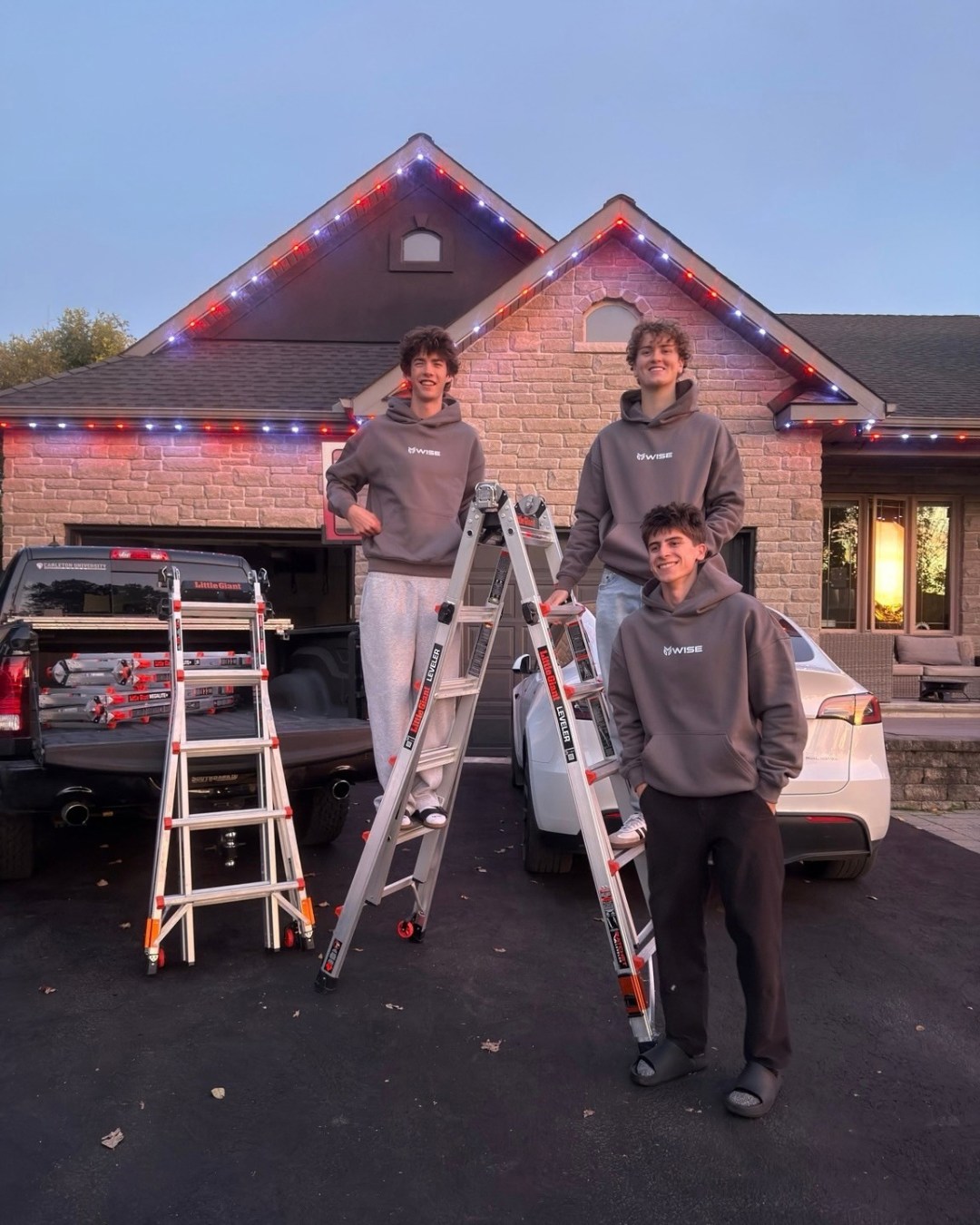 Three Wise Outdoor Lighting crew members with installation ladders and work vehicles in front of a completed red and white roofline project